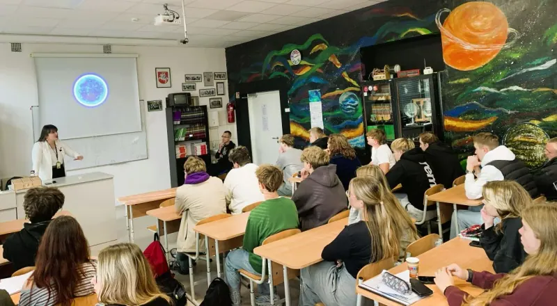 Stoviu prie lentos klasėje su kosmoso piešiniais ant sienų, ekrane – mėlyna sfera, mokiniai klausosi