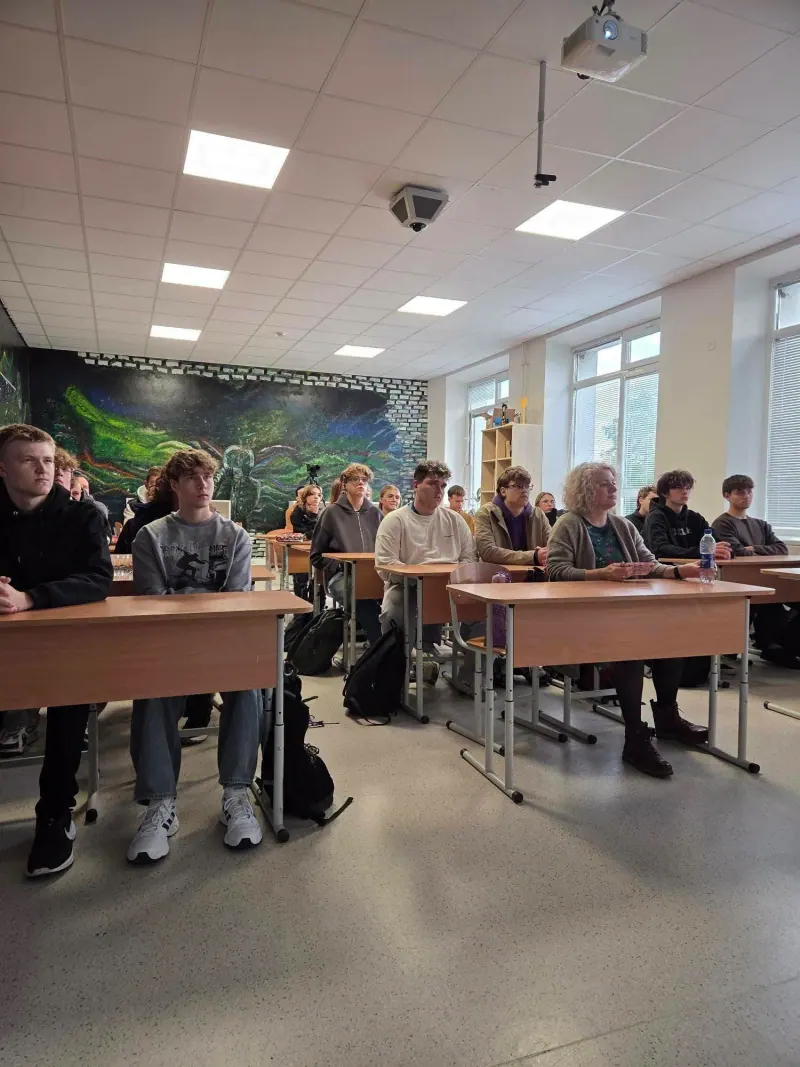 Gimnazistai ir mokytoja sėdi suoluose ir klausosi paskaitos