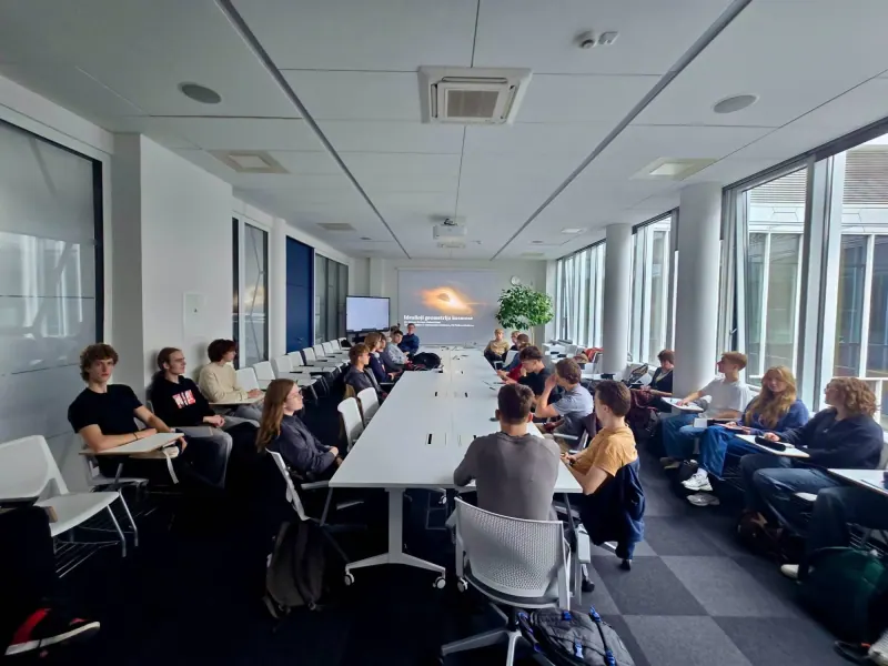 A long table in an FTMC hall with school students seated around it; at the far end a screen shows the lecture title "Idealioji geometrija kosmose" (Ideal Geometry in Space) and an image of a black hole