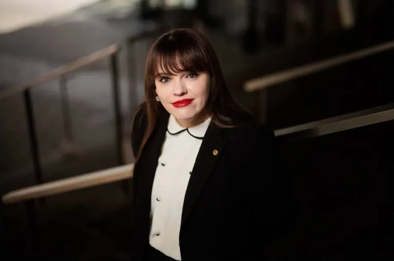 Mažena Mackoit-Sinkevičienė juodu švarku ir balta palaidine stovi prie laiptų turėklų