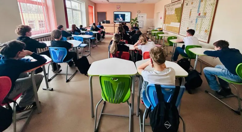 Mokiniai sėdi suoluose ir žiūri į ekraną klasės priekyje